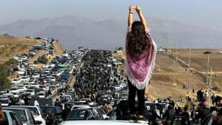 A young woman without a hijab stands on a car as a huge crowd walks towards the Aichi cemetery in Saqqez, Iran, to visit Mahsa Amini's grave on 26 October 2022