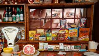 A woman looks through the window of a miniature shop that is filled with miniature products