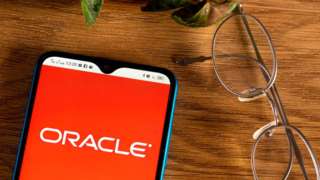 The Oracle logo is seen on an Android smartphone, on a table next to spectacles and a plant in this staged product photo