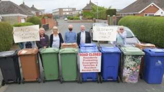 People lined up on a street with wheelie bins