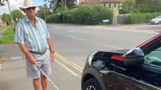Mr Fletcher using his mobility cane to negotiate a car parked on a pavement