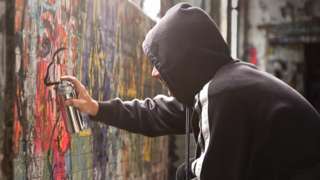 Stock image of a man with a hood spraying black paint on a wall of graffiti