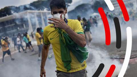 Man in mask and Brazil shirt walking towards camera with smoke behind him