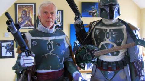 Jeremy Bulloch in Boba Fett costume standing alongside a Boba Fett figure, with Star Wars images on a wall in the background