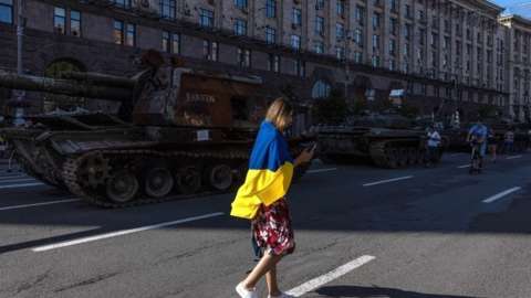 A woman with Ukrainian flag walks past Russian armoured military vehicles that were captured in fighting by the Ukrainian army, Kyiv, 24 August 2022