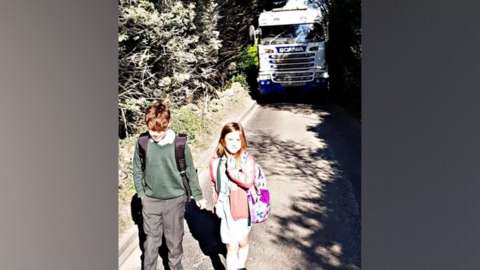 two children walking home from school with a large lorry behind them on the road