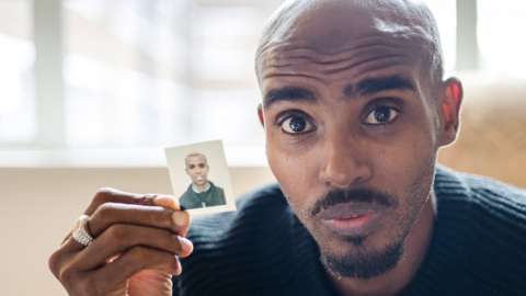 Sir Mo Farah pictured looking into the camera while holding a passport photo of himself as a child