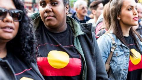 Three Aboriginal women wearing t-shirts with the Aboriginal flag on it take part in a rally against the closure of Aboriginal communities in Melbourne in 2015