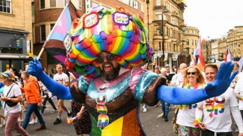 A bearded man wearing a rainbow dress parades through the city