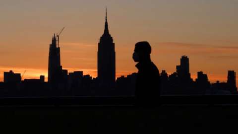 A person wearing a mask walks along the Hudson River as the sun rises behind the Empire State Building in New York City on May 14, 2020 as seen from Hoboken, NJ.