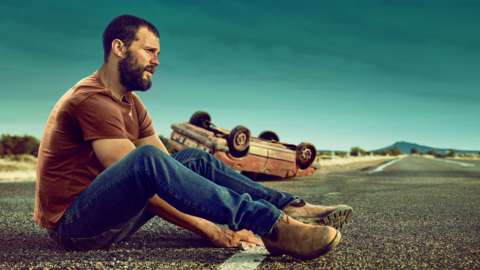 A man sat dazed at an empty roadside; with a car turned upside down behind him.