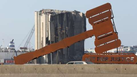An installation depicting a judge's gavel and saying "act for justice", on a wall opposite destroyed grain silos at Beirut's port (24 January 2023)