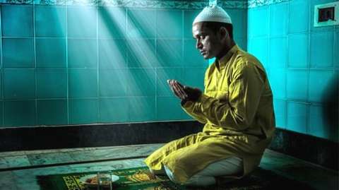 Young Muslim man praying, with folded hands. He is in traditional Indian Attire and sports a skull cap on his head.