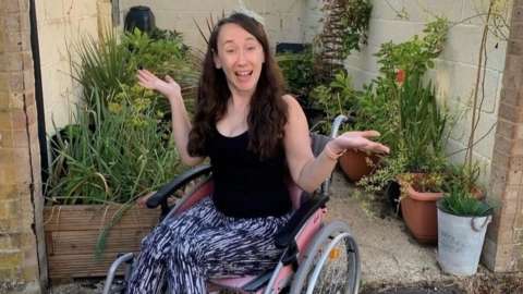 Cat Ray sitting in her wheelchair. She is wearing a black top and patterned trousers