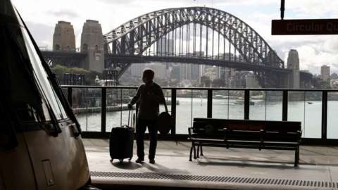 Silhouetted picture of a masked passenger on a train platform in front of the Sydney Harbour Bridge