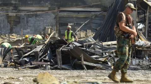 An armed Ukrainian serviceman (R) stands guard as workers (rear) clean the rubble of the destroyed Amstor shopping mall in Kremenchuk, central Ukraine