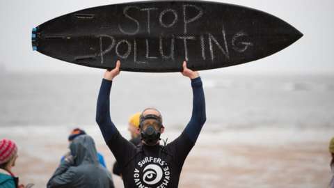 A man in a gas mask holds a surfboard up with the slogan stop polluting written on it