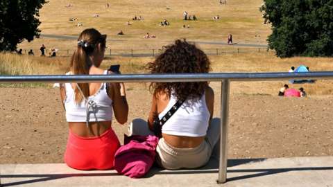 People look out toward the Old Royal Naval College, and the Canary Wharf financial district, past the sun-scorched grass in Greenwich Park, south east London on 16 July 2022