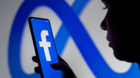 A woman holds a smartphone with Facebook's logo displayed against a background of Meta's logo