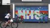 Мужчина в маске проезжает на велосипеде мимо граффити-фрески NHS в Халле