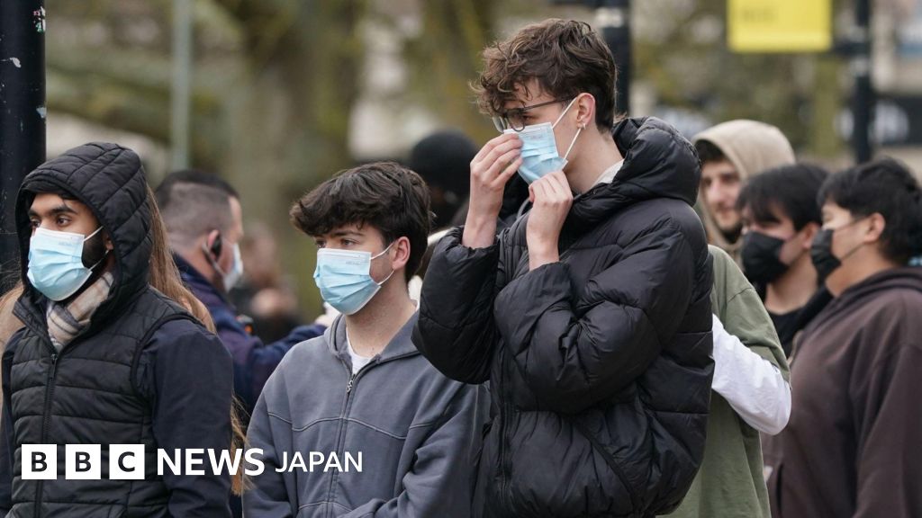 木々が植わっている屋外に大勢の若者が立っている。手前のピントがっている3人はマスクを着用している