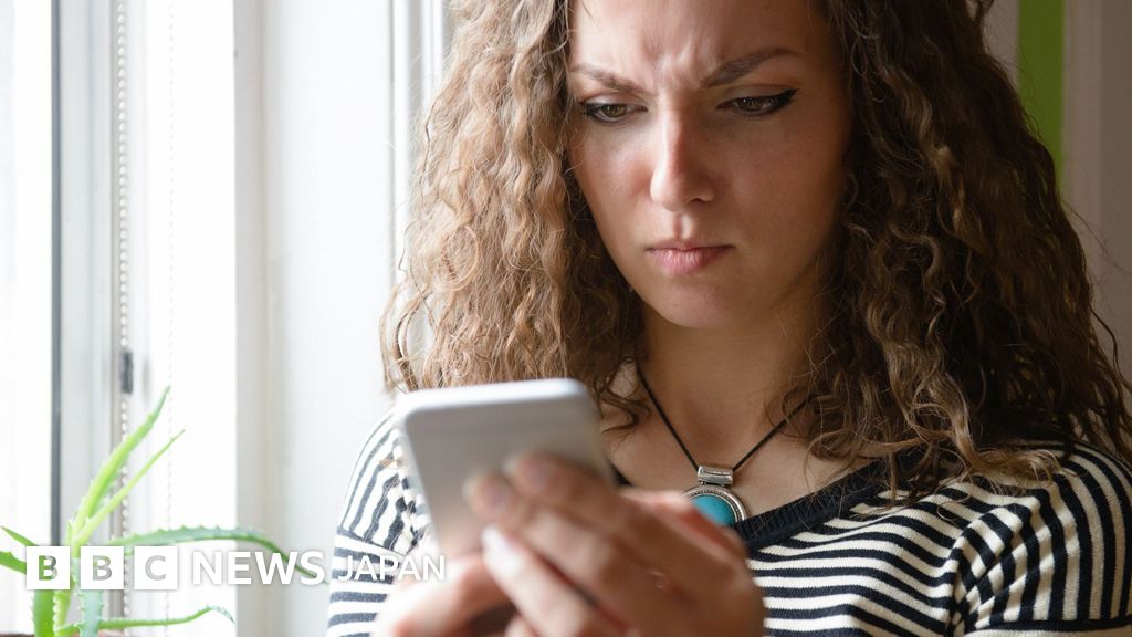 窓際にいる女性の胸から上の写真。女性は顔をしかめながら、手に持った白いスマートフォンを見ている。紺と白のボーダーのトップスを着て、黒い紐と丸い大きなターコイズでできたネックレスを着けている。窓際にはアロエの葉が見える