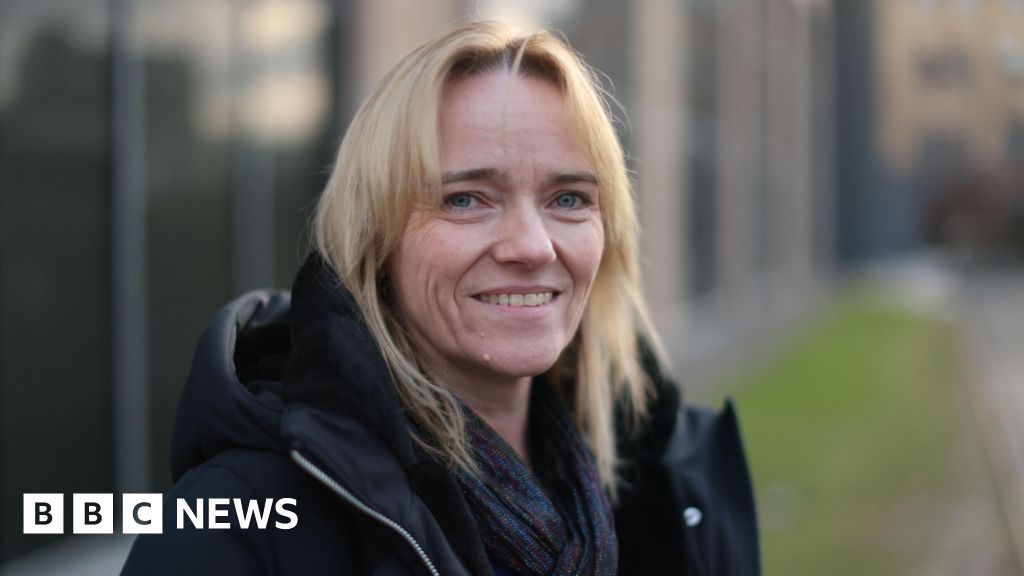 A woman with blond hair, wearing a dark jacket and smiling into the camera while standing outside an out of focus building