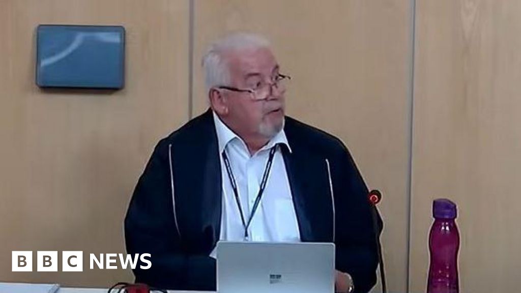 A man sitting at a table in a council meeting room. He is looking to his left. He has white hair and a white goatee and is wearing glasses. He is wearing an unzipped blue jacket over a white shirt. His laptop is open in front of him and there is a purp...