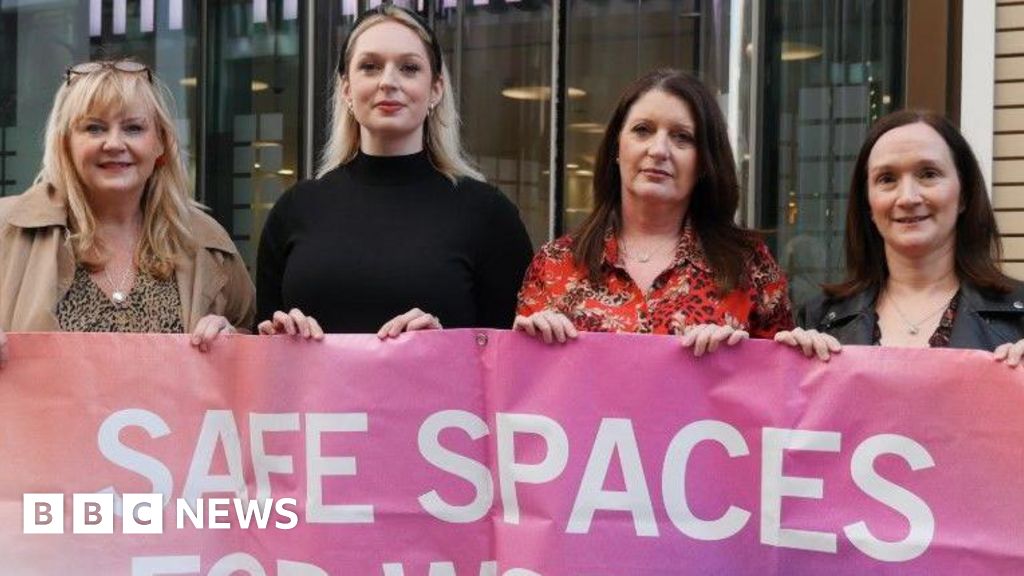 Four women stand outside an office building holding a pink and purple banner with the slogan "Safe spaces for women" written in large white letters.