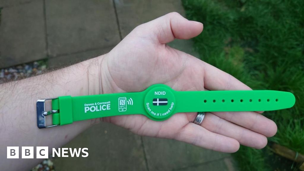 A hand laying flat. There is a wristband lying on the palm of the hand. It is green, has a Cornish flag in the centre and says "NDID" and "Scan me if I need help" in the centre. On the left strap it says "Devon and Cornwall Police" with a picture of a...