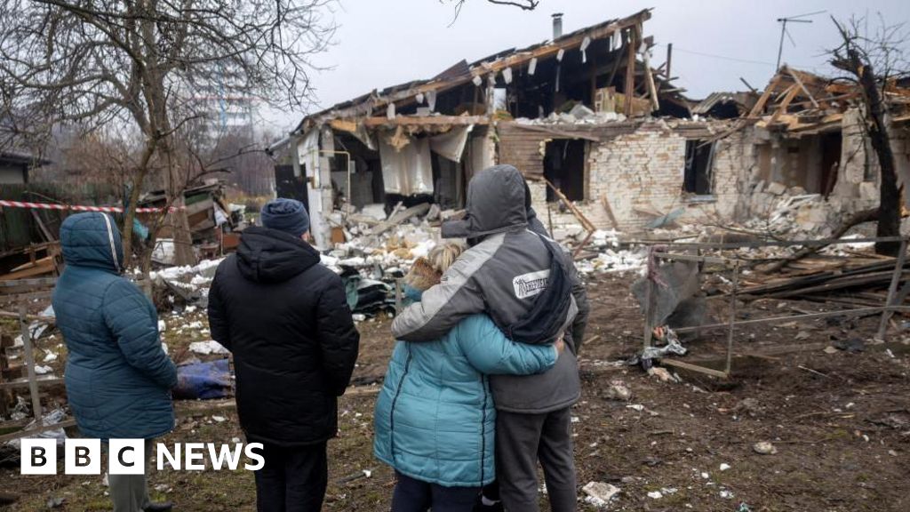 Four people in winter jackets look at ruined house.
