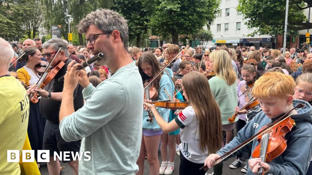 In the foreground, a man plays the flute. Behind him is a young boy concentrating as he plays the fiddle. In the background hundreds of other musicians play their instruments.