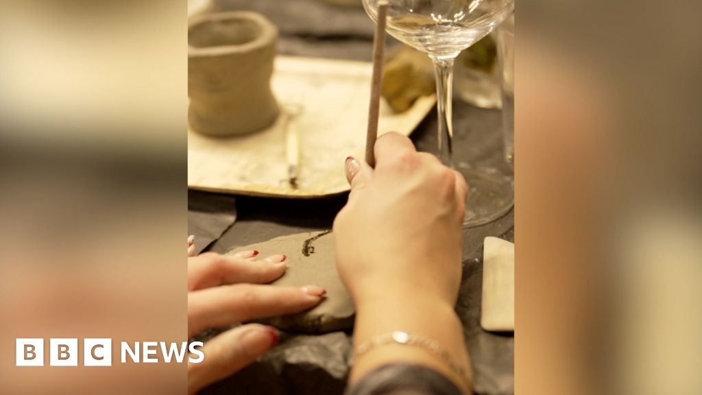 A pair of hands using a wooden tool to make marks in a piece of clay. A wine glass sits on the table