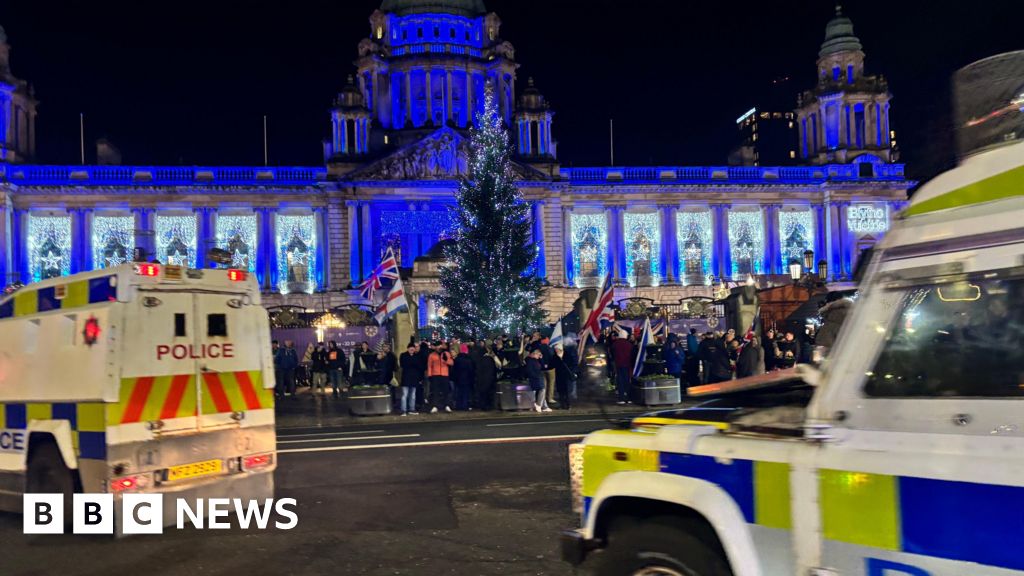 A blue lit Belfast City Hall. There is a Christmas tree in front and people carrying Union Jack flags have gathered at the gates. Two police land rovers are on the road.