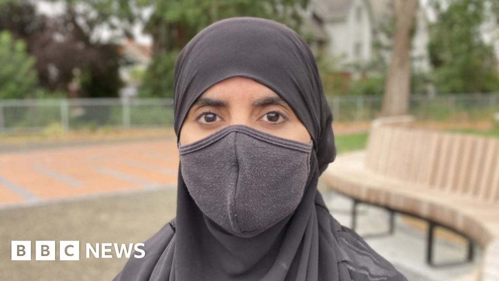 A woman wearing a black hijab and black face mask stands in front of a grassy area of a residential street.