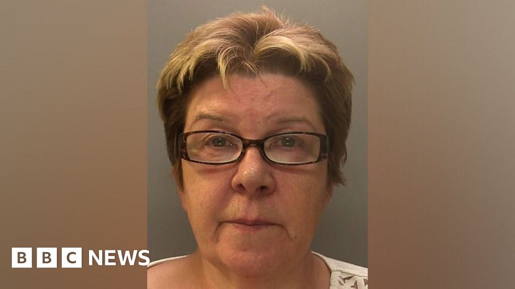 A police mugshot of an older lady - she has cropped ginger hair, black glasses and is wearing a white top.