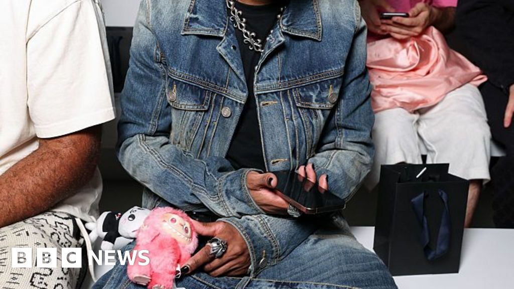 D4vid sits down, wearing a matching jean jacket and baggy pants, black shades, and carrying a pink Labubu doll