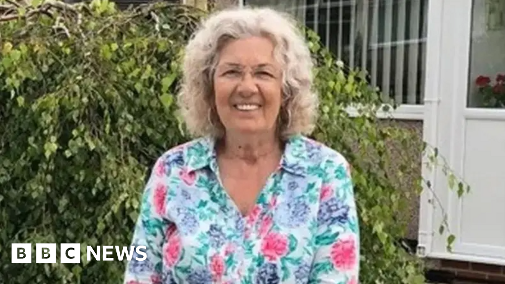 Valerie Kneale, with shoulder-length grey curly hair, wears a pink, lilac and green floral blouse as she is photographed in a garden. She is standing in front of a pebble-dashed house with a white plastic door and windows.
