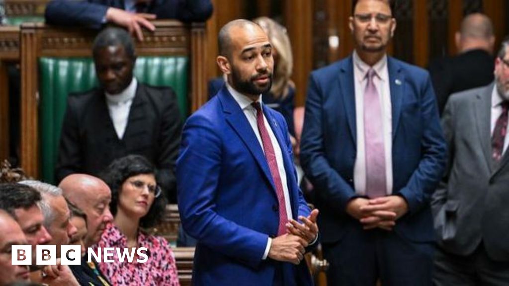 A man in a cobalt coloured suit and a red tie standing up and talking in parliament.