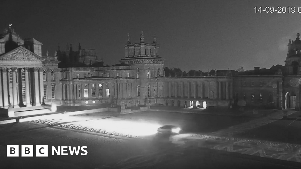 A night CCTV image of Blenheim Palace courtyard showing a car driving through it with headlights on. The ground leading up to the palace is overed in large Union flags