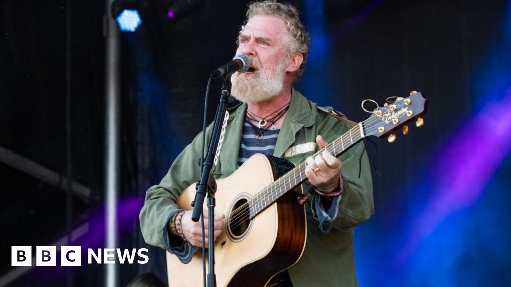 Photo: BBC Glen Hansard sings into a microphone while holding an acoustic guitar. He has a white beard and grey short hair. He is