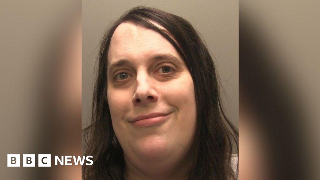 A police headshot of a trans woman with long brown hair smiling into the camera. The background is grey.