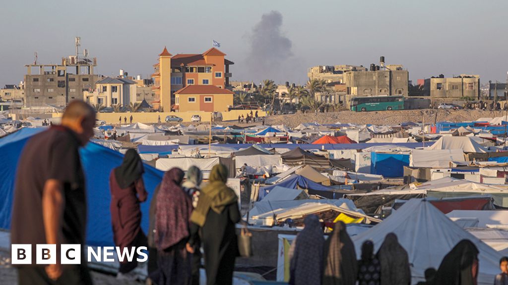 Smoke rises in the distance while people stand in front of a tent community in Rafah on 7 May