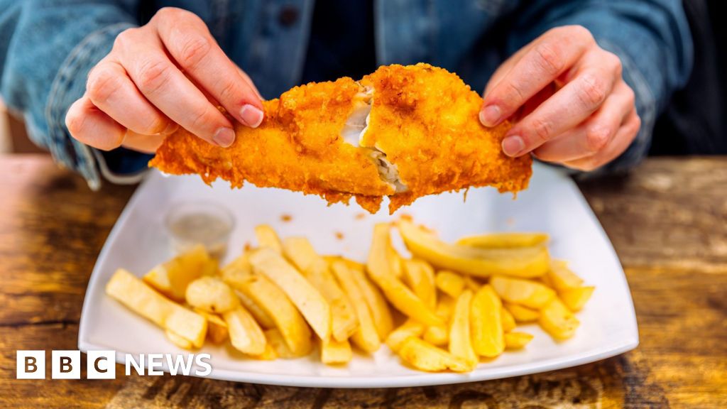 Plate of fish and chips on a wooden table, with someone holding the fish about to eat it.