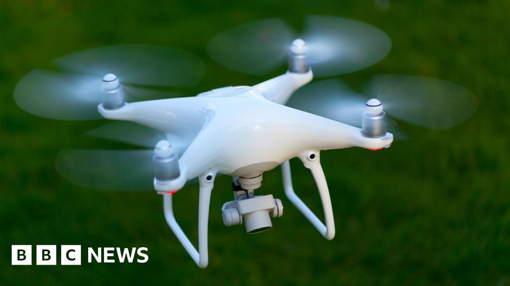 A white drone with four propellors and a camera looking down underneath it
