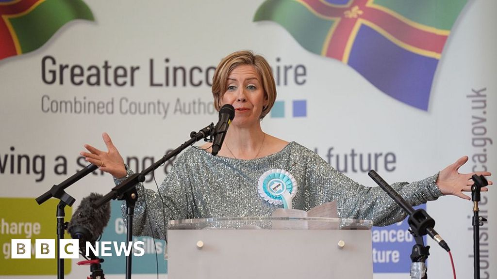 Andrea Jenkyns making a speech after winning the Lincolnshire mayor election. She is wearing a silver top with a blue Reform UK rosette and is standing behind a podium surrounded by microphones and in front of banner with Lincolnshire flags on it
