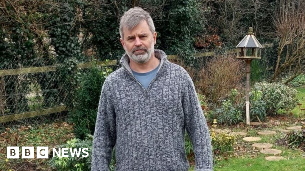 Kevin Lister, with grey hair and beard, stands in a garden wearing a grey knitted jumper