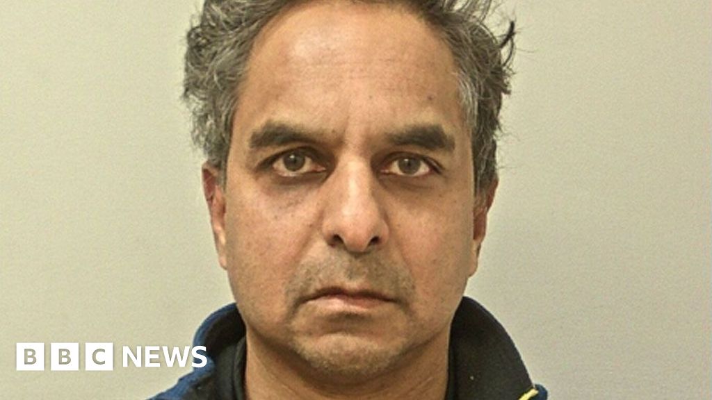 Police mugshot of Dr Amal Bose. He has short, unkempt dark hair that is greying and is looking directly at the camera, frowning slightly. He has brown eyes and bushy eyebrows.