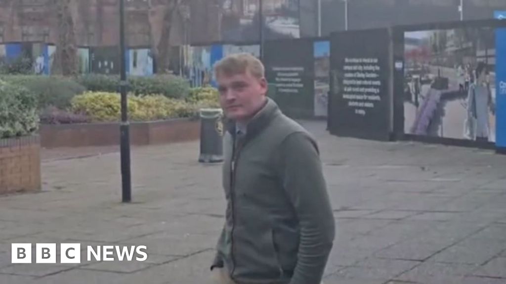 A man with short blonde hair and a grey jacked standing in a paved street with hoardings behind him