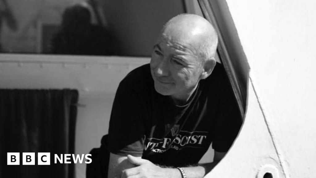 A black and white photo of Fra Hughes, a man with receding white hair, on board a boat.  He is wearing a black t-shirt with an "anti-facist" logo and a wrist band.